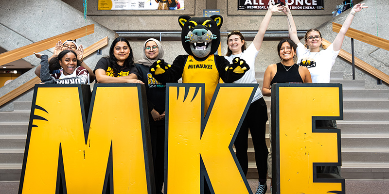 UWM Students and mascot, Pounce, smiling in UWM Union in front of tall "MKE" sign
